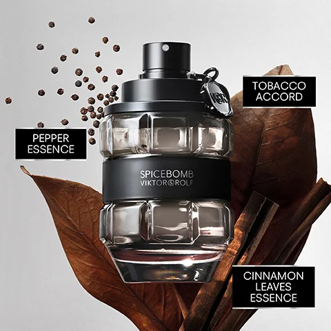 A Spicebomb Viktor&Rolf perfume bottle shaped like a grenade stands amidst peppercorns, dried leaves, and cinnamon sticks, with labels pointing to PEPPER ESSENCE, TOBACCO ACCORD, and CINNAMON LEAVES ESSENCE.